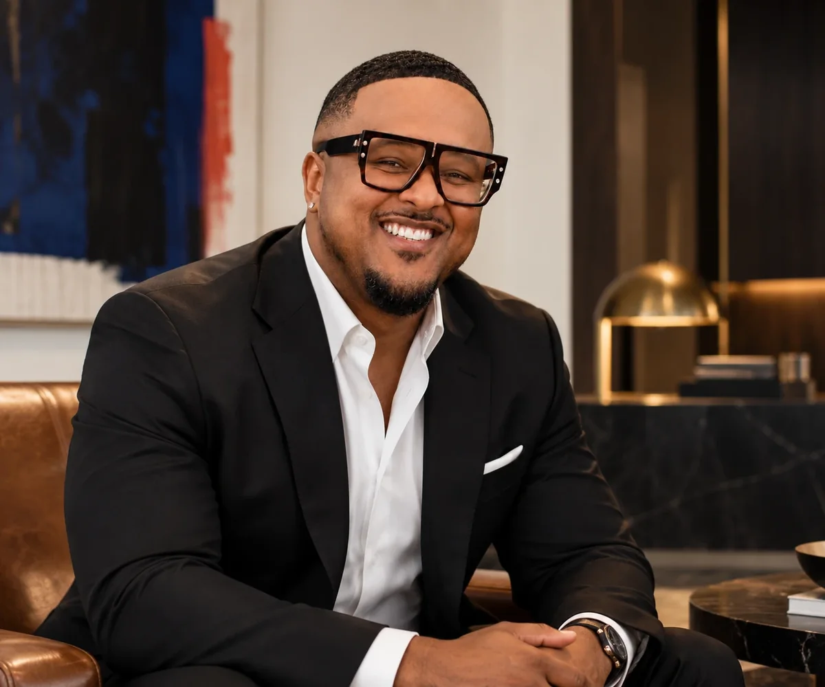 Brennon Jackson, founder of EYEGASMIC, smiling in a black jacket and white shirt in an architectural lounge.
